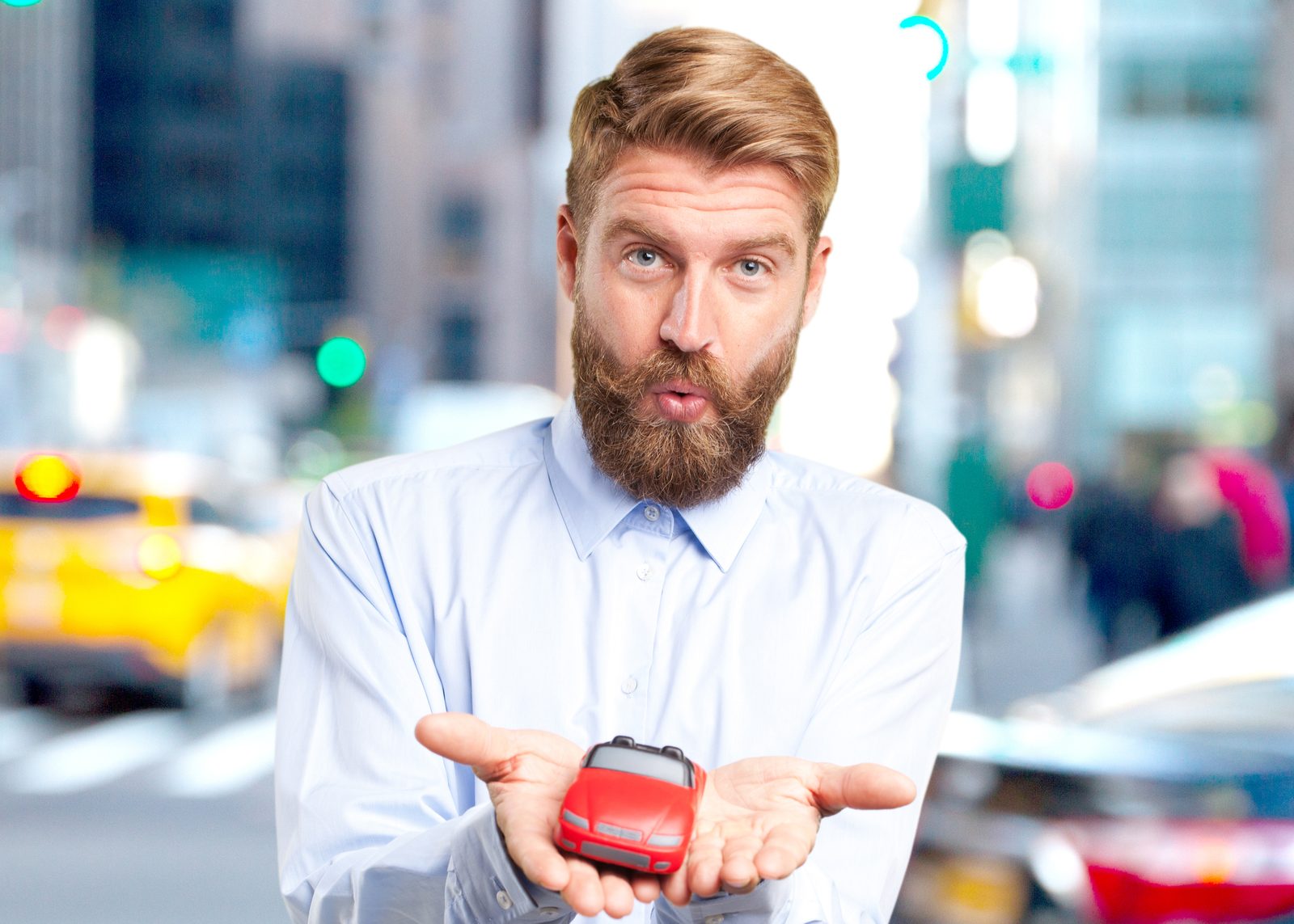 Un homme barbu en chemise tient une voiture miniature rouge dans ses mains, dans une rue